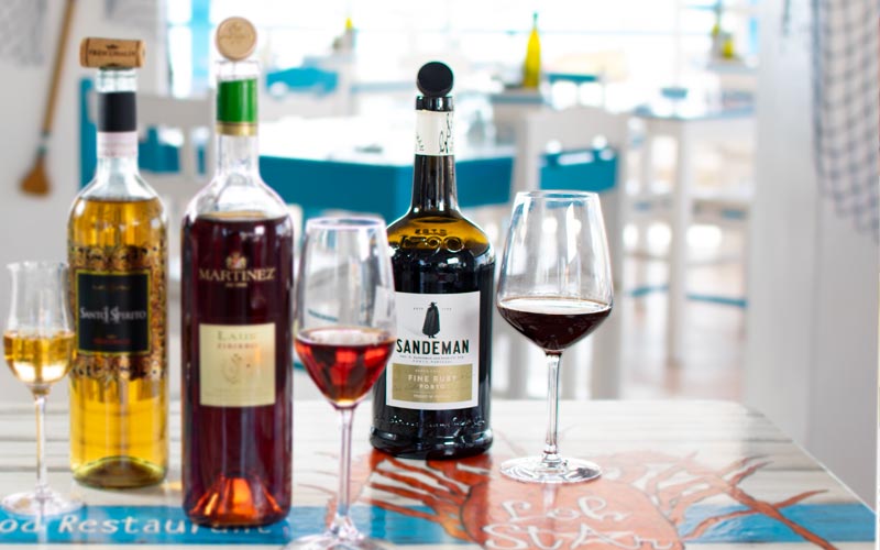 PalmBeach Tropical Restaurant | Porto Fine Ruby DOC Sandeman <small>(al calice)</small> | Vol. 19,5% / Touriga Franca, Tinta Roriz, Tinta Amarela, Tinta Barroca, Tinto Cão / DOC Porto / Portugal, Douro / Colore rosso ambrato chiaro, corpo leggero e intenso. Al naso, profumi di vaniglia e frutta secca evoluta. In bocca, si sviluppa un’elegante armonia di sapori di frutta rossa fresca, combinata con la complessità della maturazione in legno e un finale persistente.